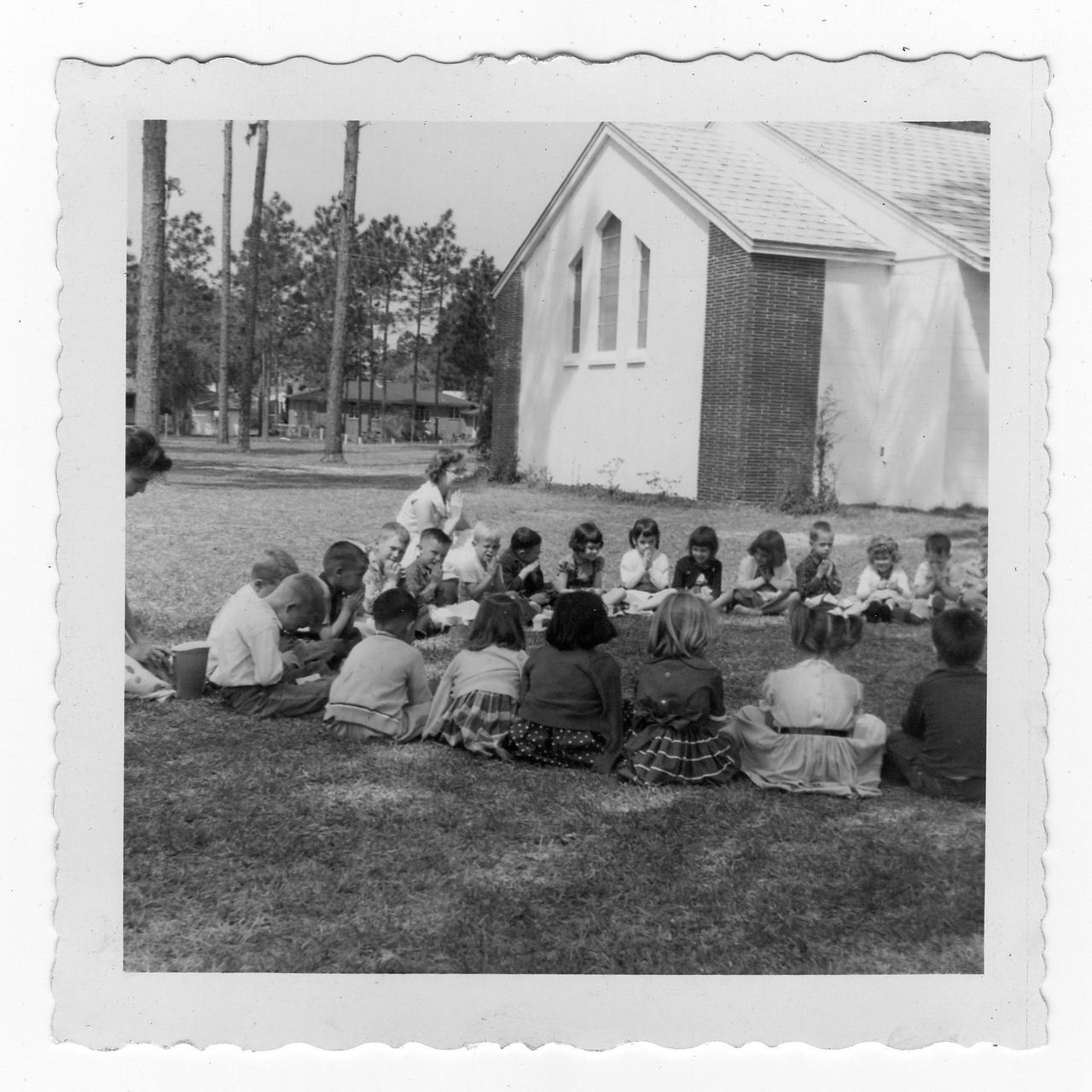 Children seated in a circle on grass, listening to a teacher in front of a small white church building.