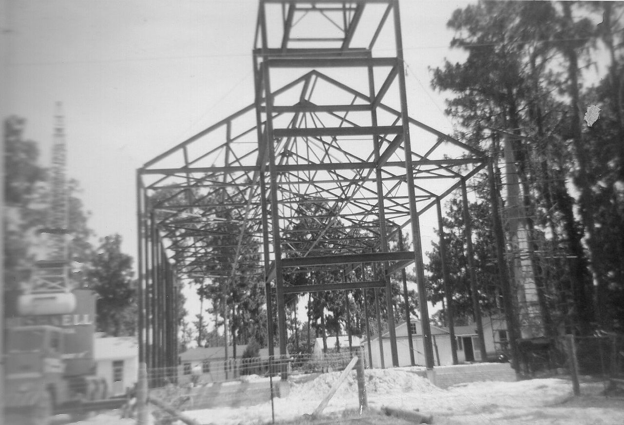 Construction of a large, skeletal metal structure, possibly a building or enclosure, amidst trees.
