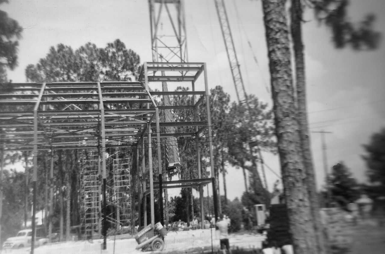 Construction of a metal structure with a tall tower. Men are working on the scaffolding in a wooded area.