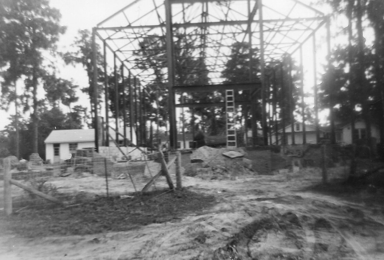 Construction site with a metal frame rising amidst trees, dirt, and small buildings.