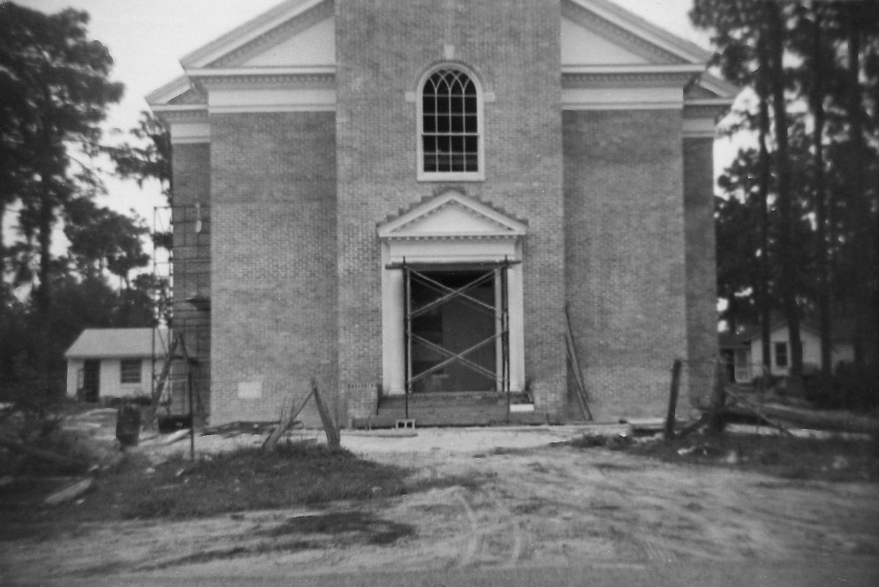 A large, light-colored building with scaffolding around the entrance; small building to the left, trees in the background.