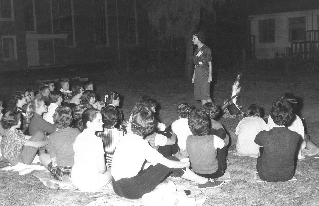 Group of people seated on the ground listening to a standing person outside at night.