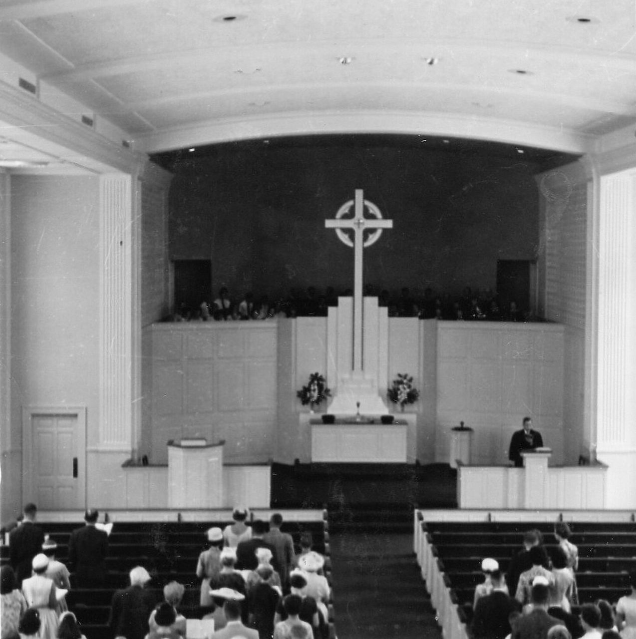 Interior of a church with people seated in pews, a large cross on a stage, and a speaker at a lectern.