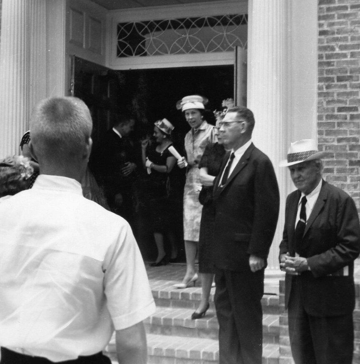 People exiting a building with columns. Man in white shirt faces the viewer. Others wear suits and hats.