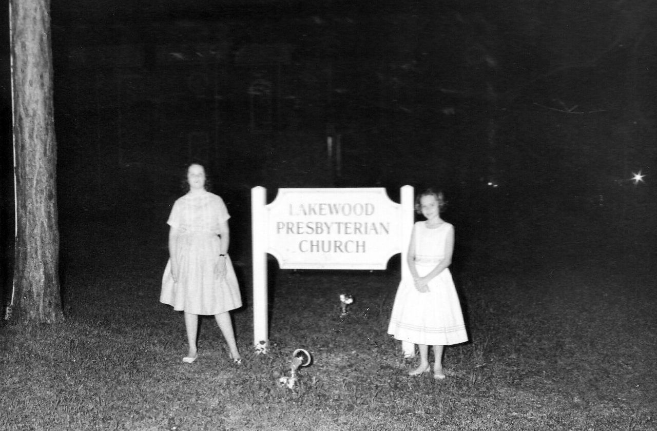 Two women stand by a sign for Lakewood Presbyterian Church at night.