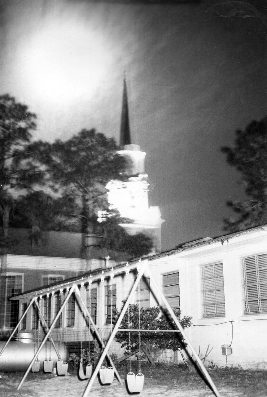 Black and white photo of a church steeple glowing above a playground with swings.