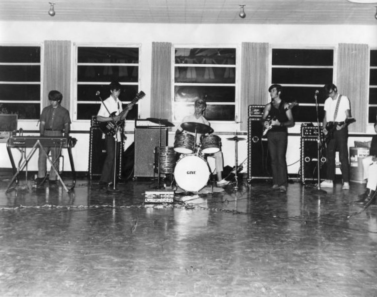 Band performing on a stage. Musicians play guitar, drums, and a keyboard. Venue has windows and a wood floor.