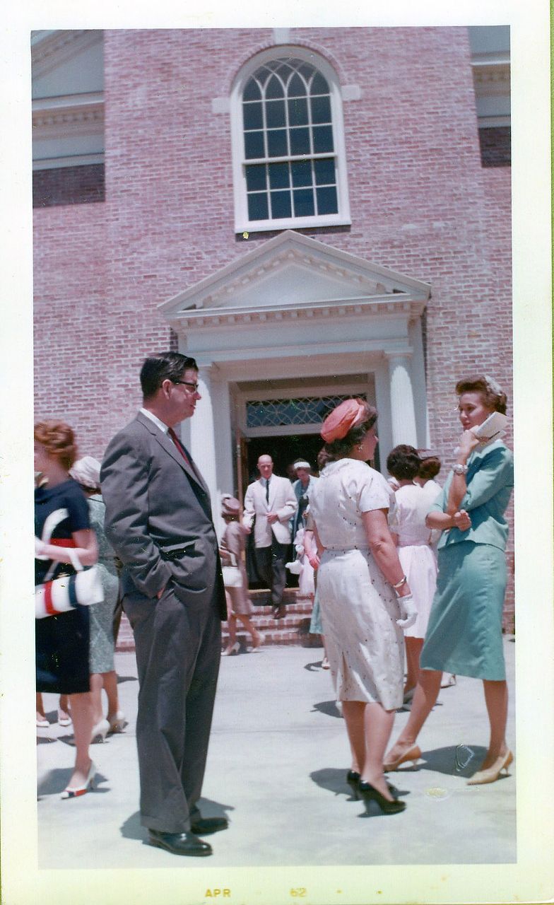 People gather outside a brick building with a white arched window. Man in a suit, women in dresses, sunny day.
