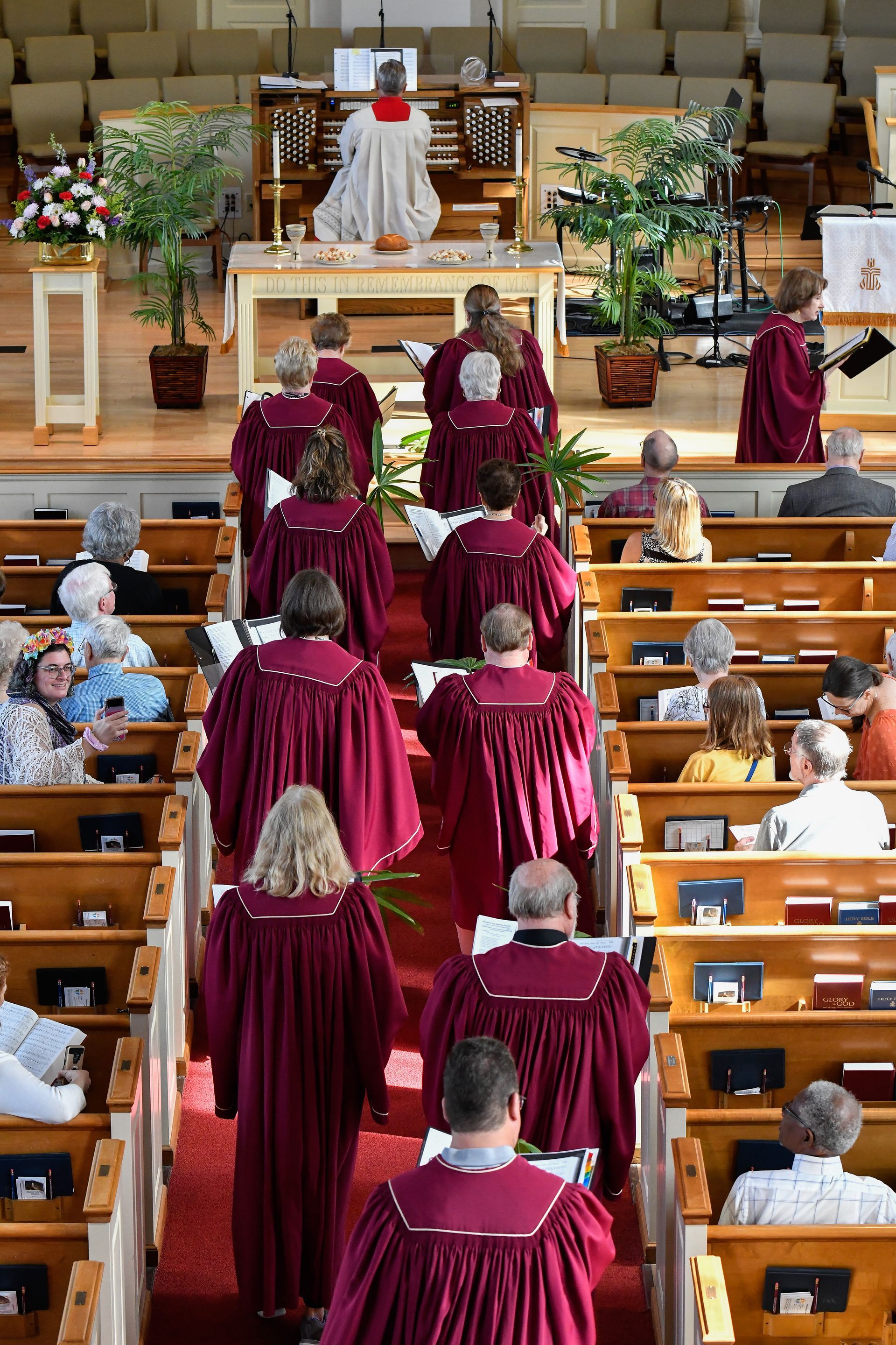 Choir in burgundy robes walks down a church aisle towards a podium; audience seated in pews.