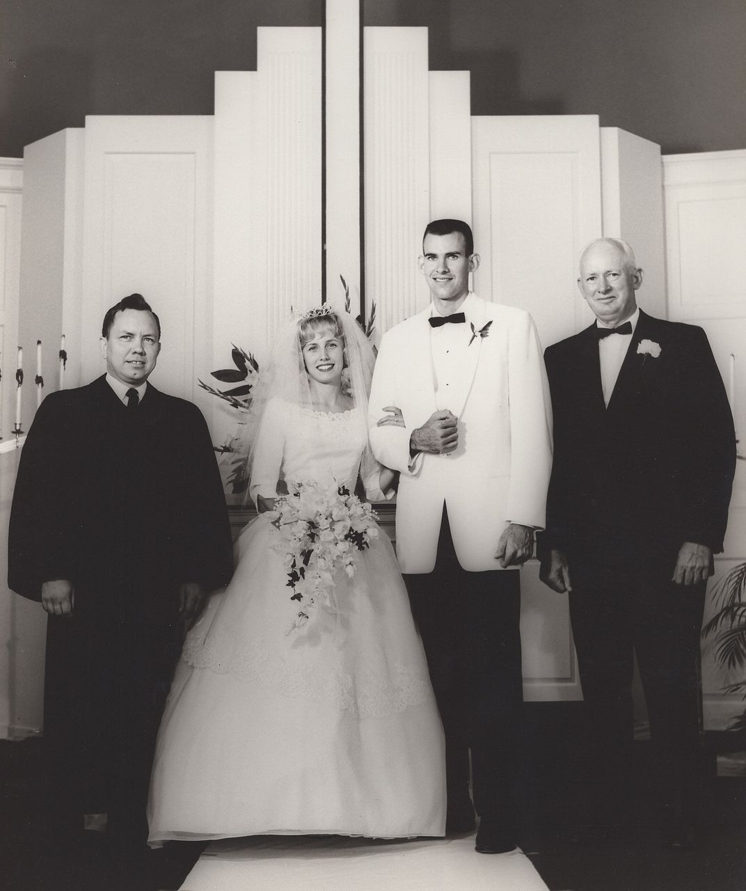 Wedding photo: bride, groom, officiant, and another man pose in front of a decorative wall.
