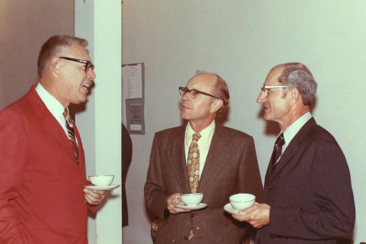 Three men in suits converse, holding teacups. Indoors, likely an office setting.