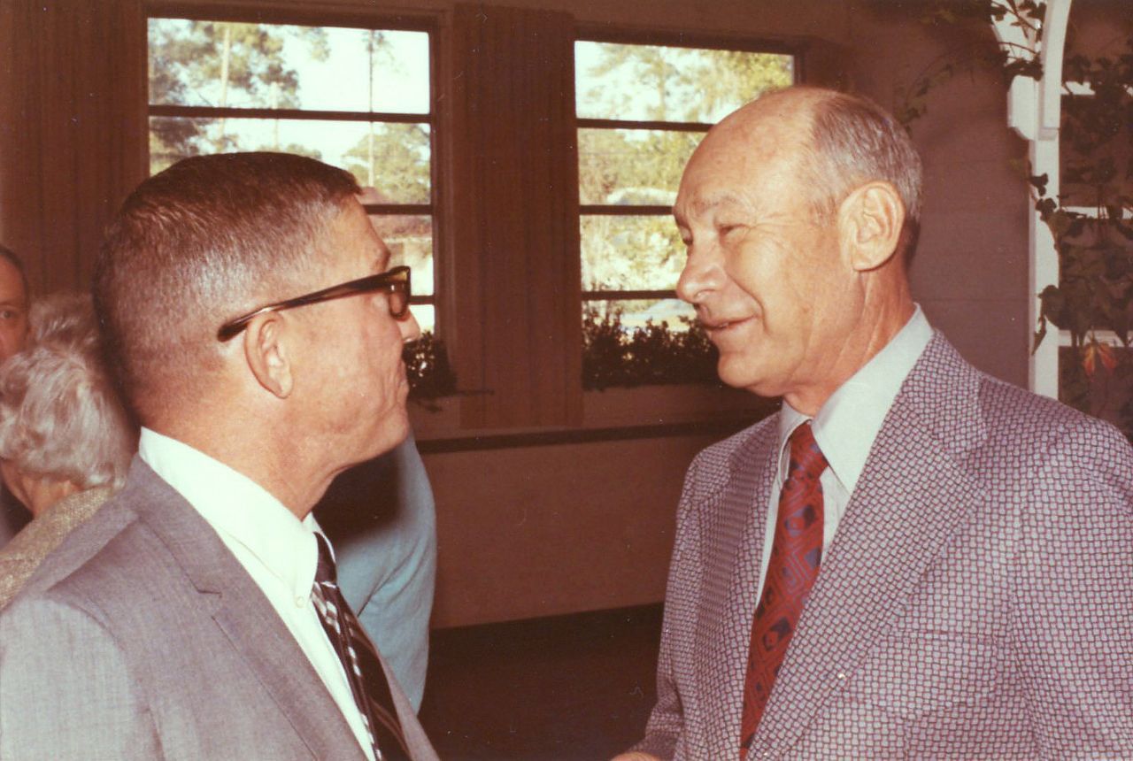 Two men in suits conversing, possibly indoors. One wears glasses, the other smiles. Soft lighting.