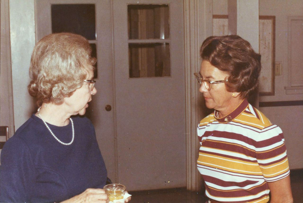 Two women in a room, one in a striped shirt, the other in a blue top and pearl necklace, talking.
