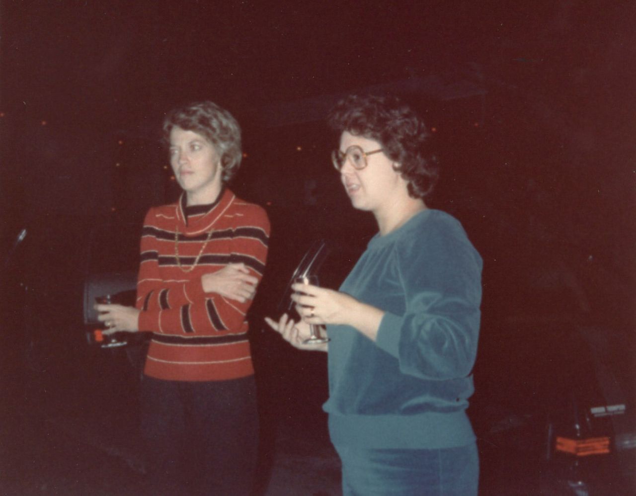 Two women holding drinks stand in a dark room. One in striped red sweater, other in teal sweatshirt, both talking.