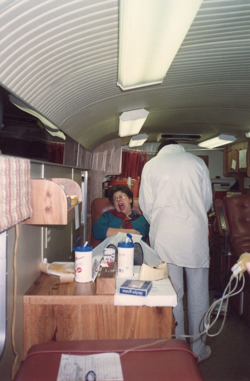 Inside a blood donation bus, a person reclines as a healthcare worker attends to them. Red chairs, wood paneling.