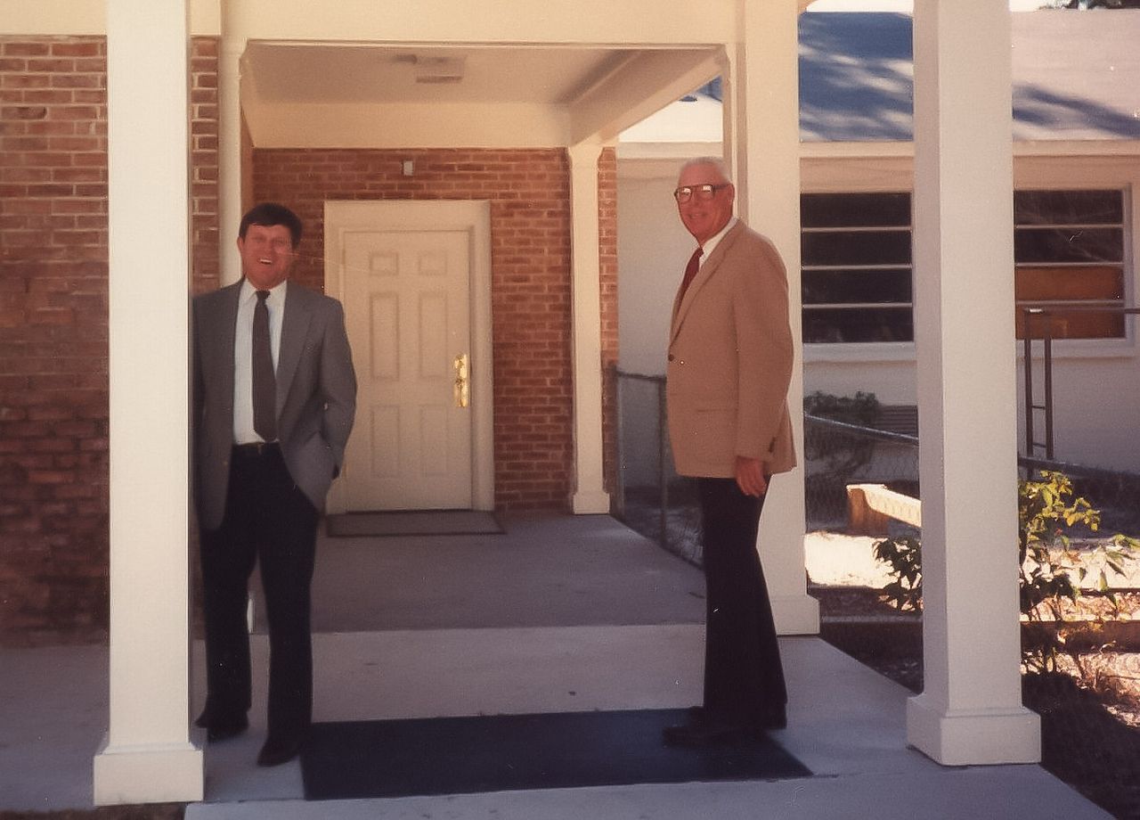 Two men in suits stand outside a brick building with white columns. One man leans on a column.