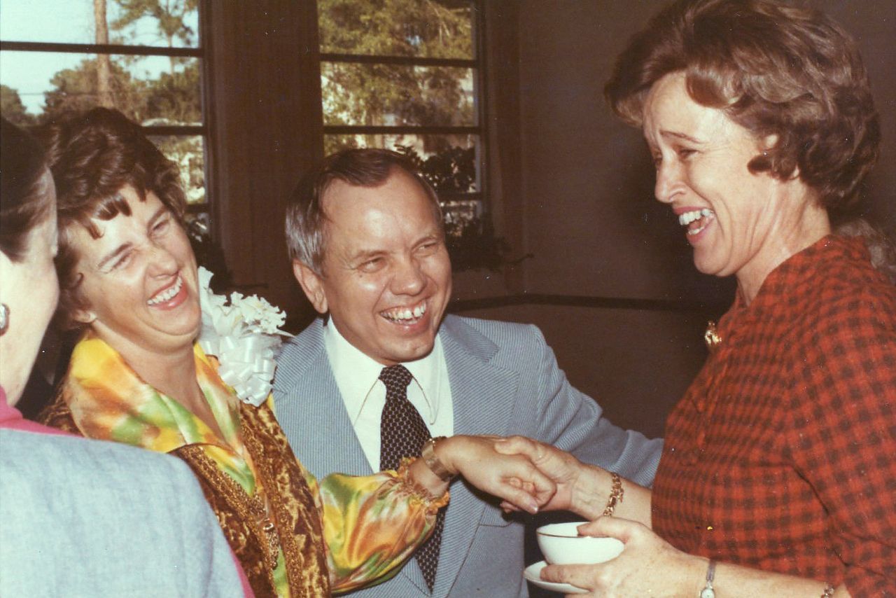 Three people laughing, shaking hands. Man in a suit, two women, one in a colorful blouse, the other in a red patterned dress.