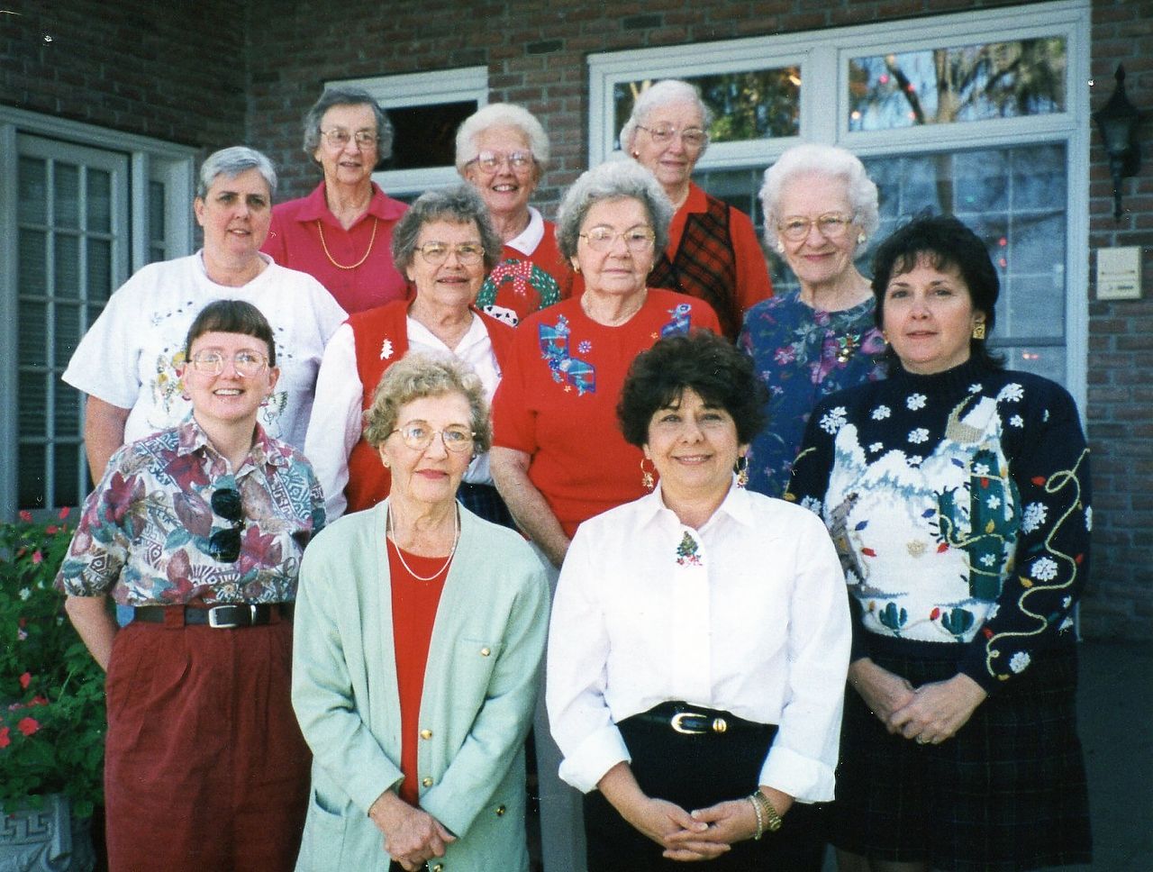 Group of women posing outside a building; some in red shirts, smiles, sunny setting.