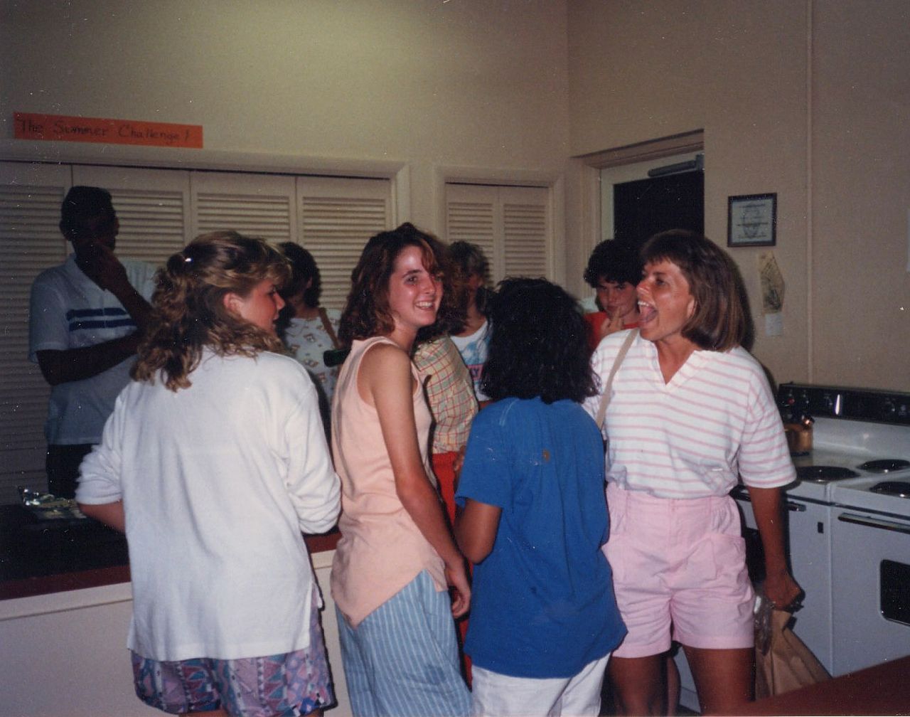Group of people gathered in a kitchen, smiling and talking. One person in pink shorts stands by stove.