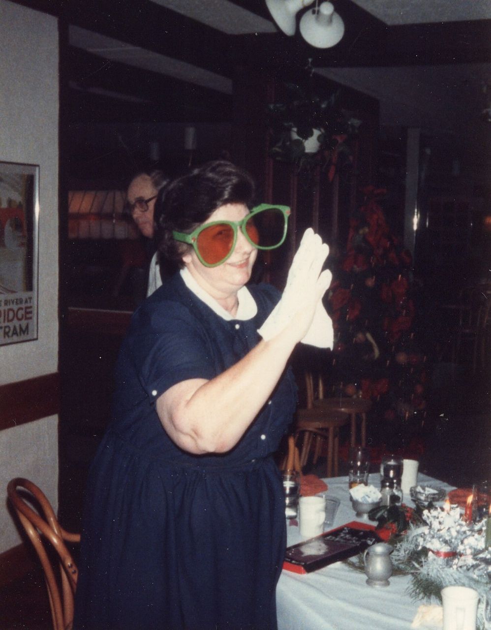 Woman wearing oversized novelty glasses and white gloves, clapping at a decorated table in a dimly lit room.