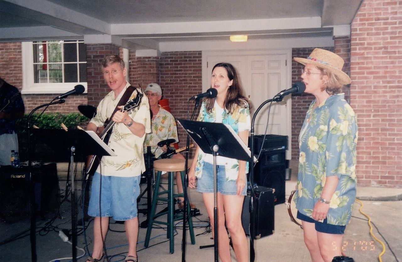 Band playing outdoors, man on guitar, two women singing, and a man on drums, wearing Hawaiian shirts.