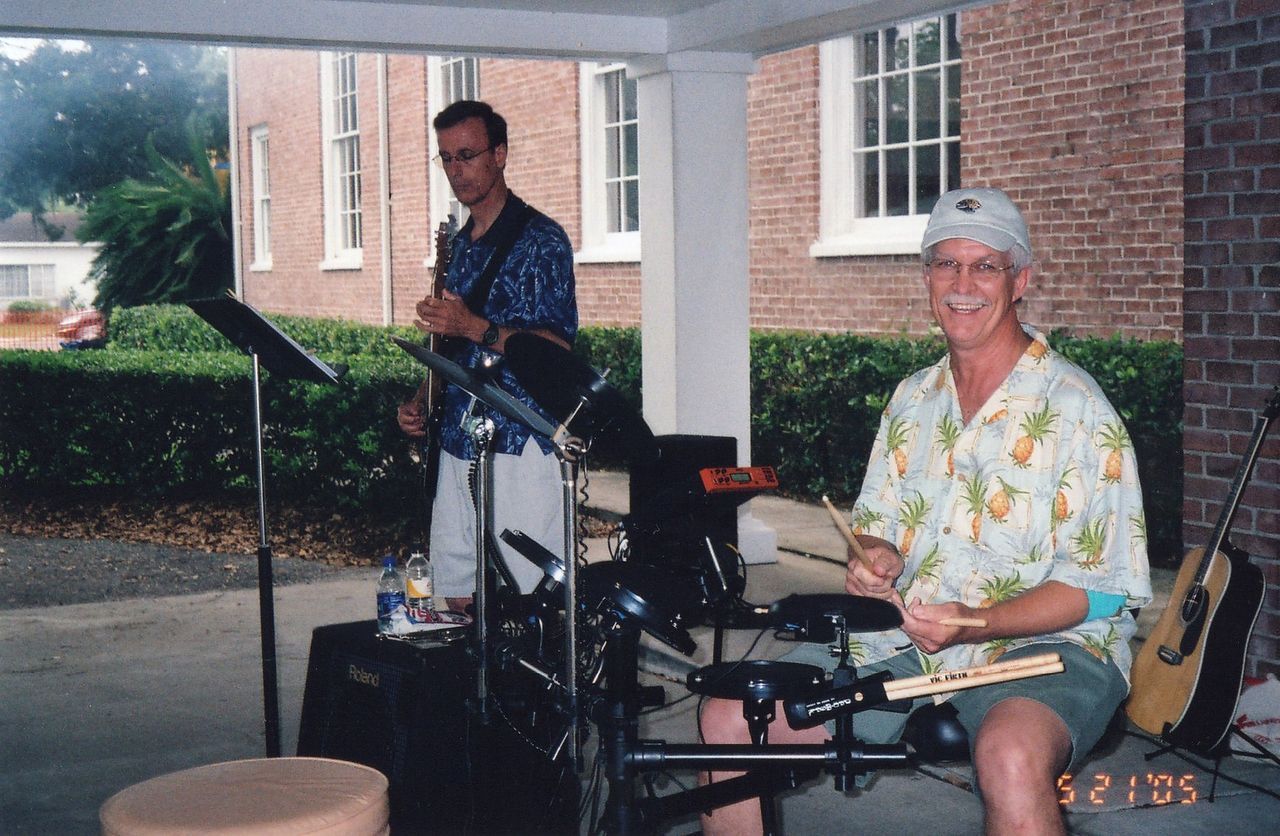 Two men playing music outdoors; one on drums, the other with a stringed instrument, under a porch.