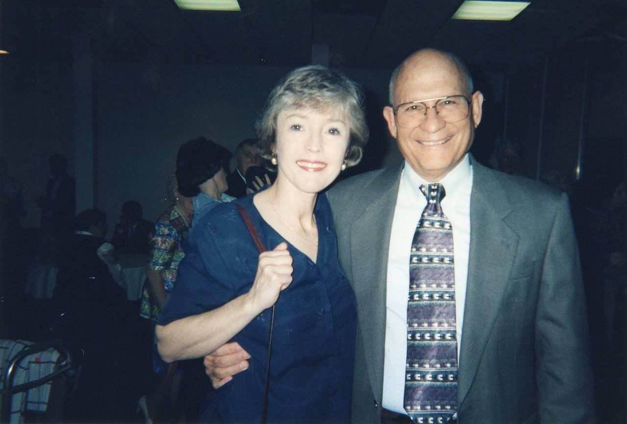 Man in suit and woman in blue shirt, smiling, posing together indoors.