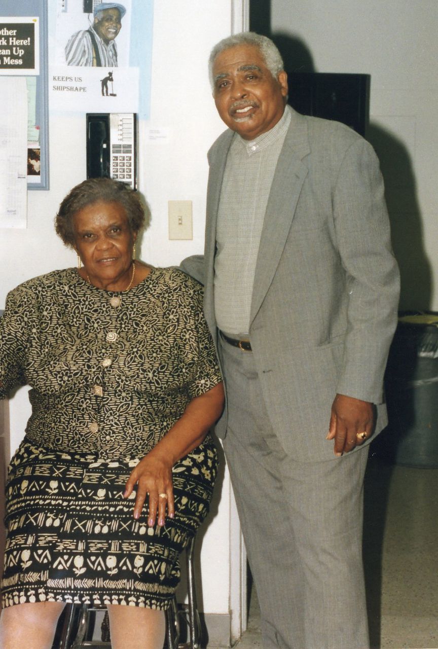 Woman seated, wearing patterned dress, with man in suit, indoors.
