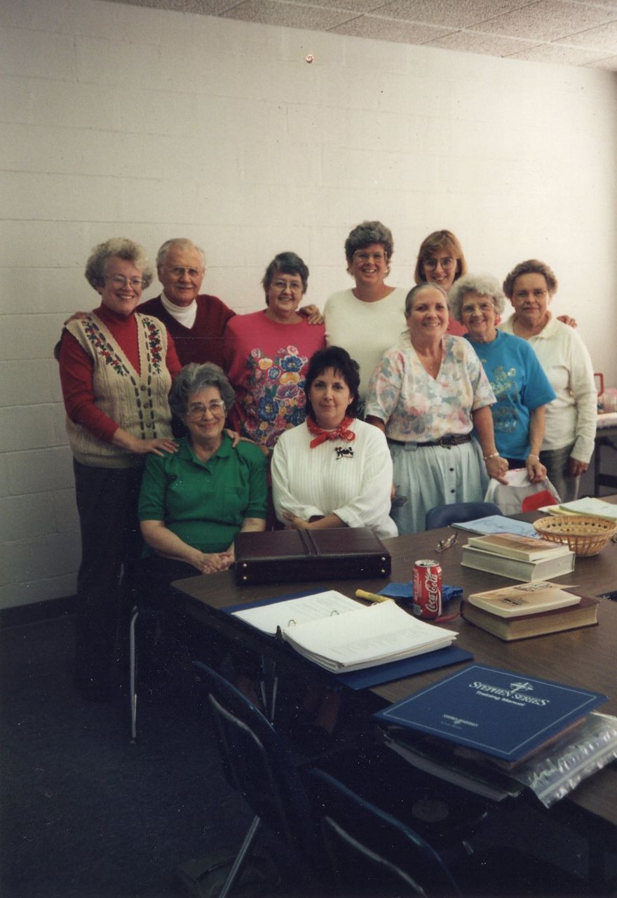 Group of people gathered around a table. Some are smiling, indoors, with papers and a soda on the table.
