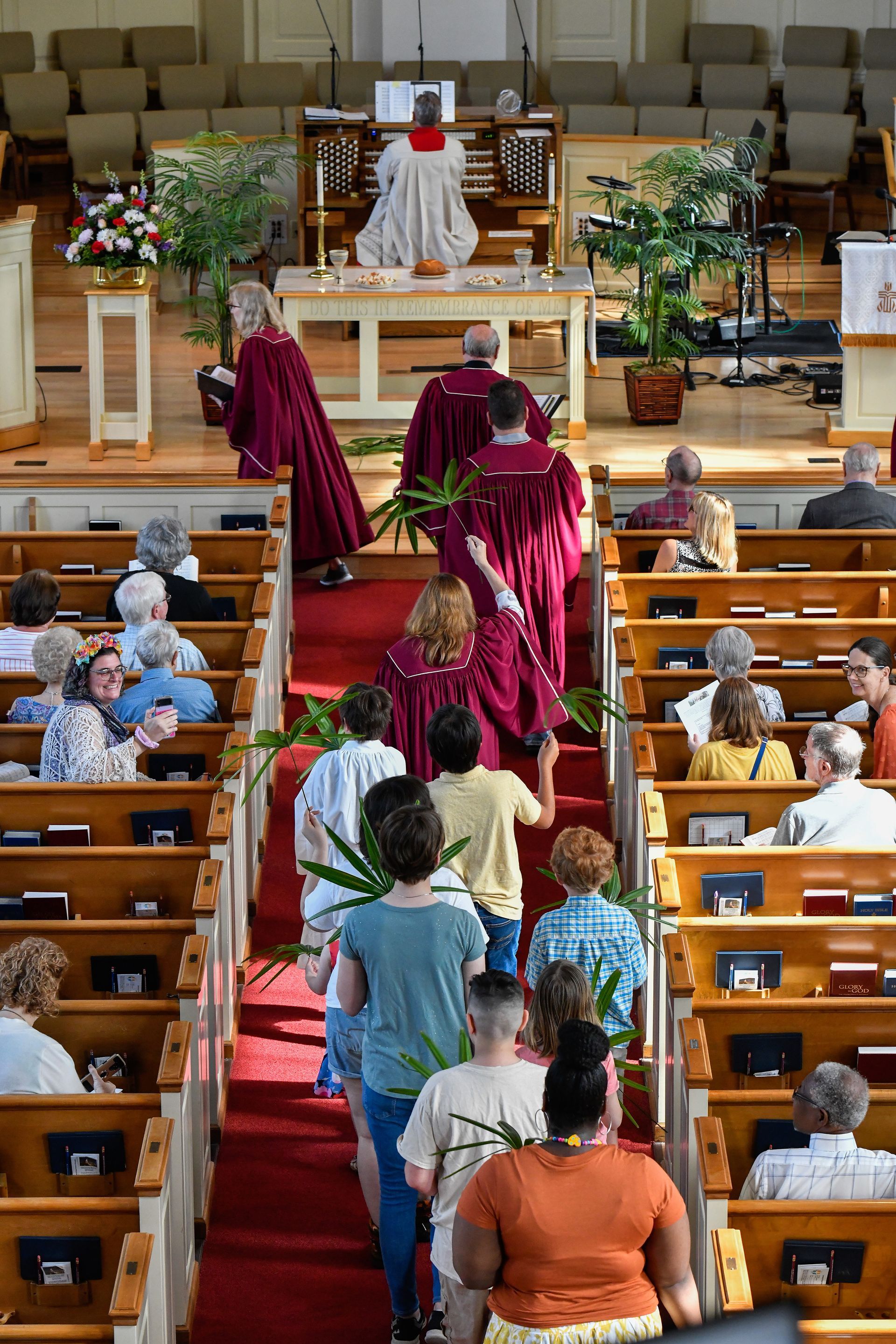 People walking up church aisle, holding palm fronds, with choir in maroon robes.