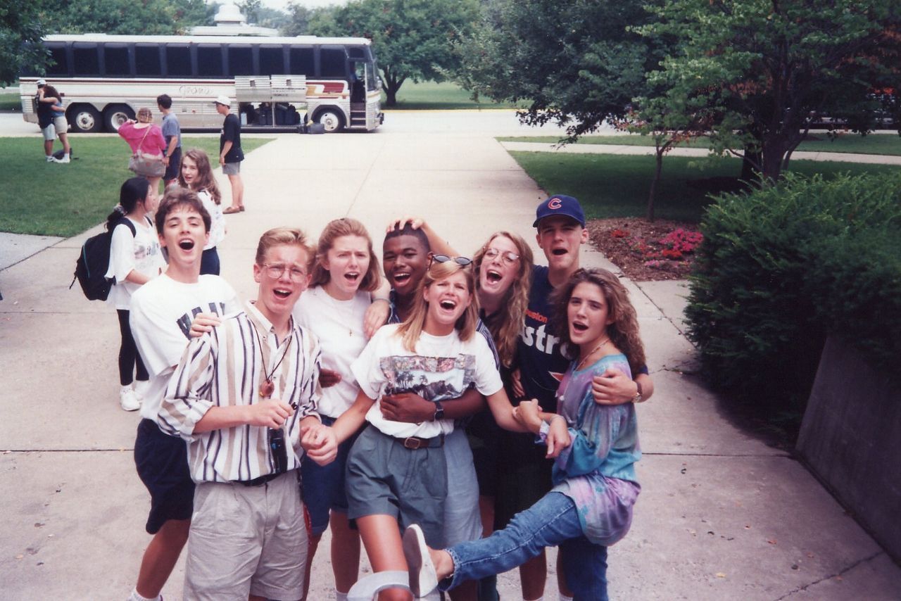 Group of people smiling and posing in front of a bus on a paved area, trees in the background.