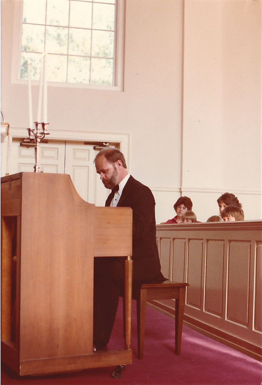 Man playing organ in church; candles on altar. Two people watch from pew.