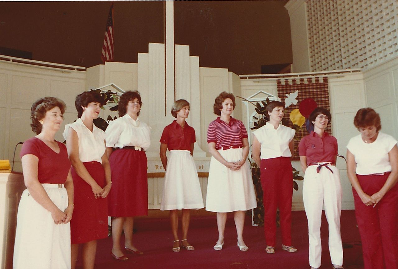 Group of women in red and white outfits stand on a stage. American flag in background.