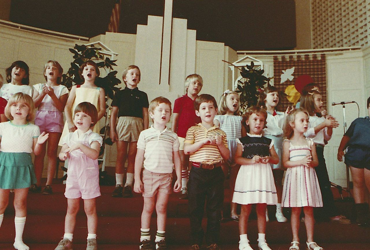 Children singing on a stage, some clapping, indoors. Brick-like backdrop.