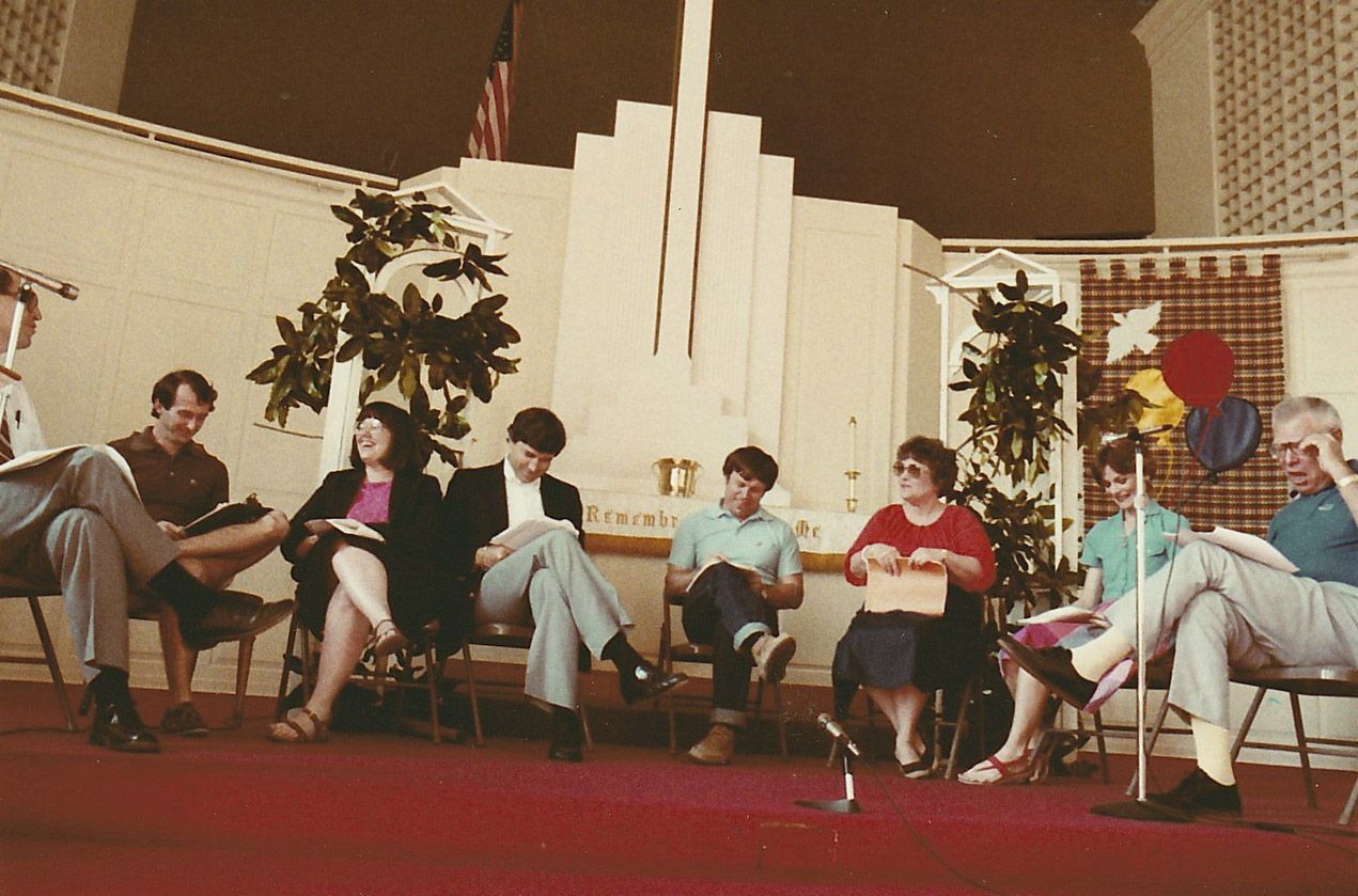 Group of people seated, reading from papers, on a stage with plants and backdrop decorations.