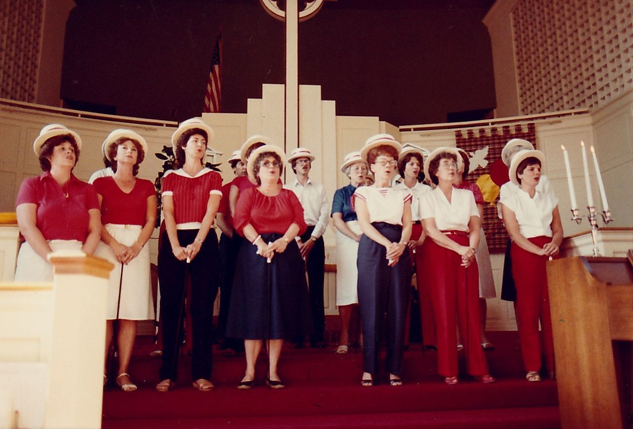 Choir in a church singing, wearing straw hats and colorful outfits, standing on a red carpet.