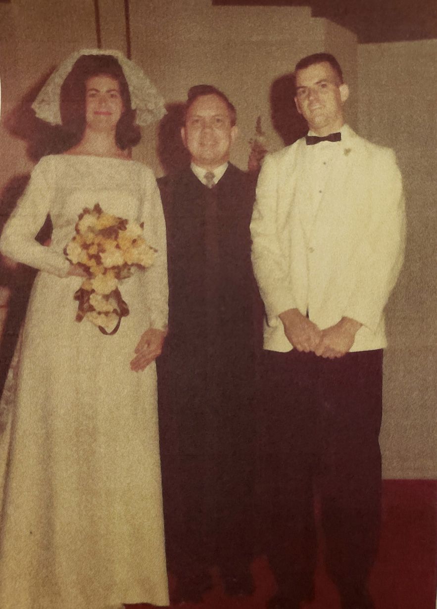 Bride and groom stand with a man in formal wear. The bride holds flowers, the groom wears a white jacket.