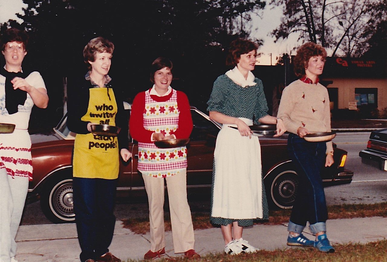 Five people in aprons holding pans stand outdoors in front of cars.