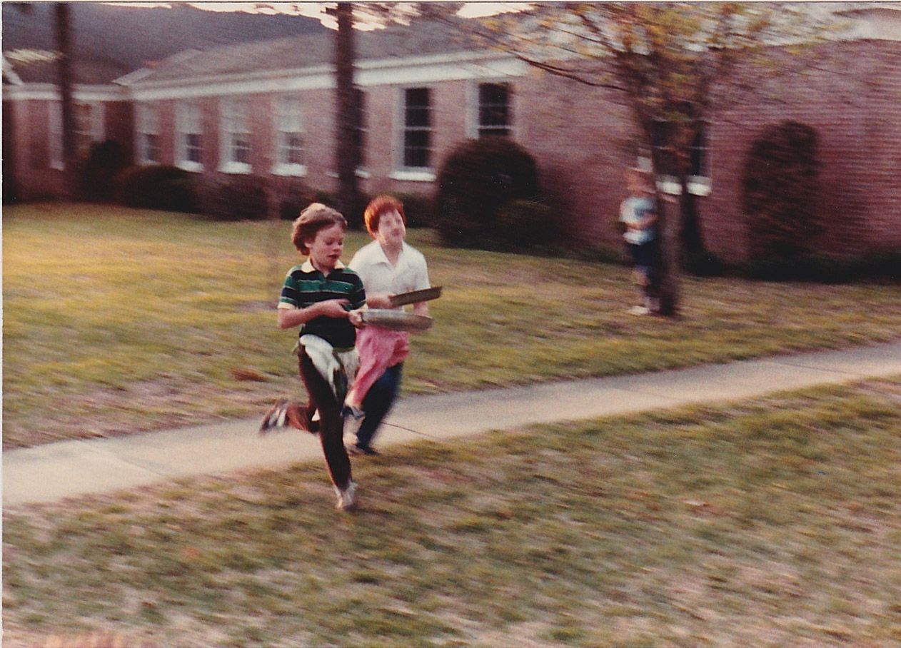 Two kids running with books in front of a brick building on a grassy lawn.