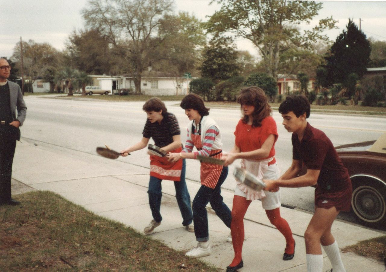 Four people flipping pancakes in a street race, wearing aprons and holding pans. An observer stands to the side.