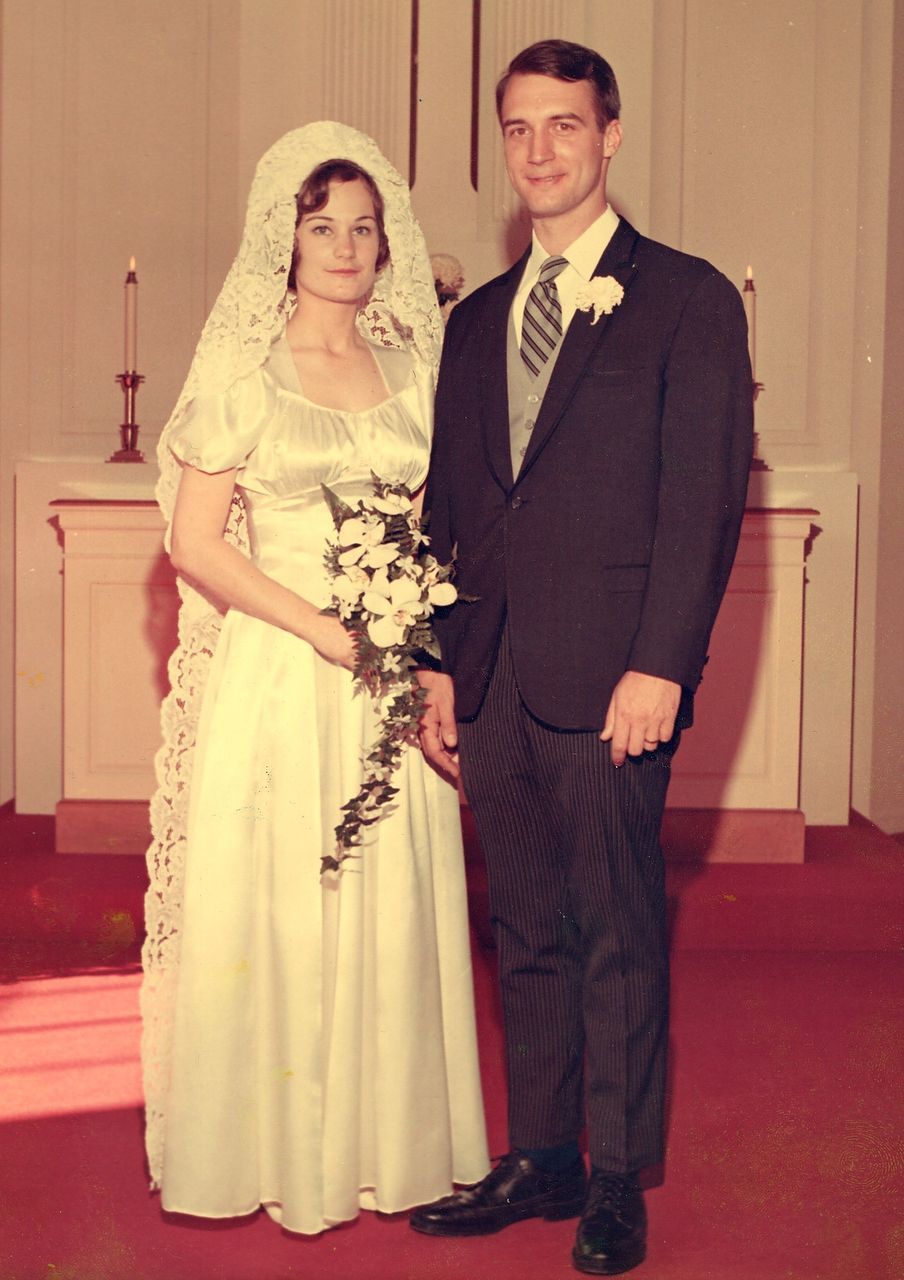 Wedding photo: Bride in white dress and veil, holding bouquet; groom in suit. Standing in a church.