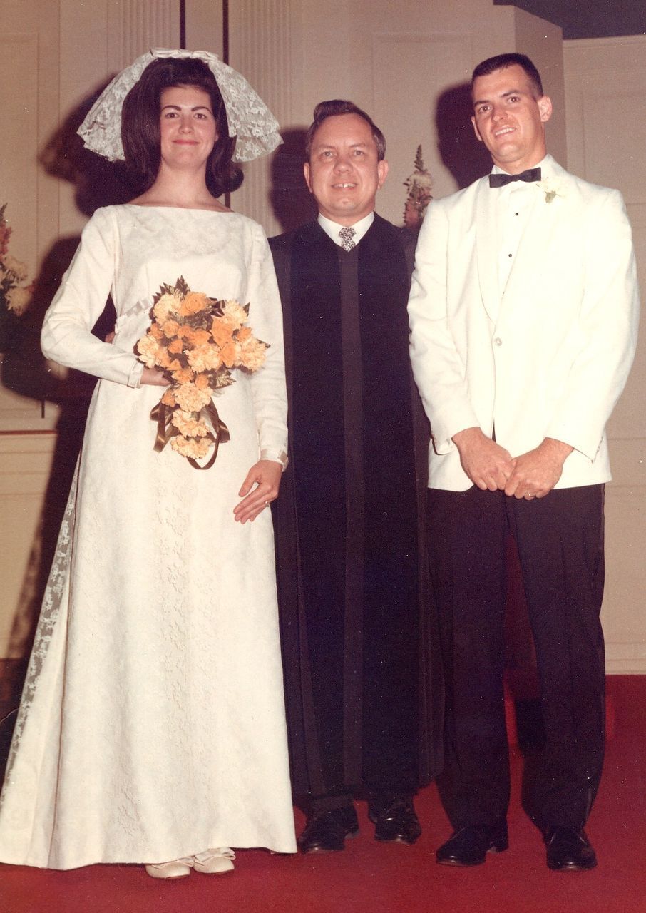 Bride and groom with officiant at wedding. Bride in white dress, groom in tux, all standing indoors.