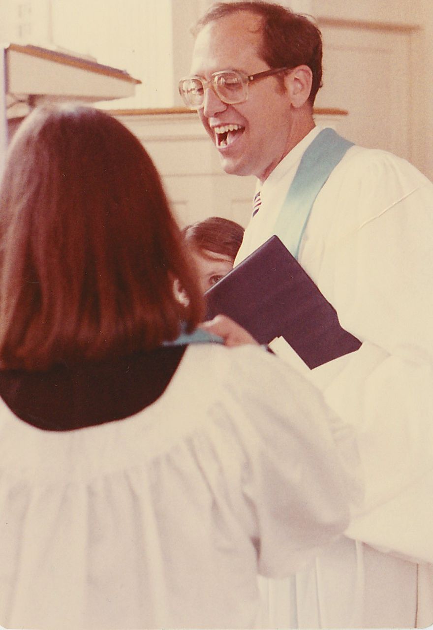 Clergy member laughing with choir member. Man in white robe, light blue stole, glasses. Interior, potentially a church.
