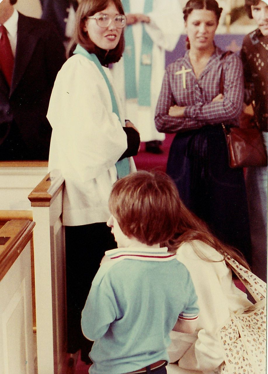 Woman in clerical collar, child, and others in church. Light-colored clothing and a blue stole are visible.