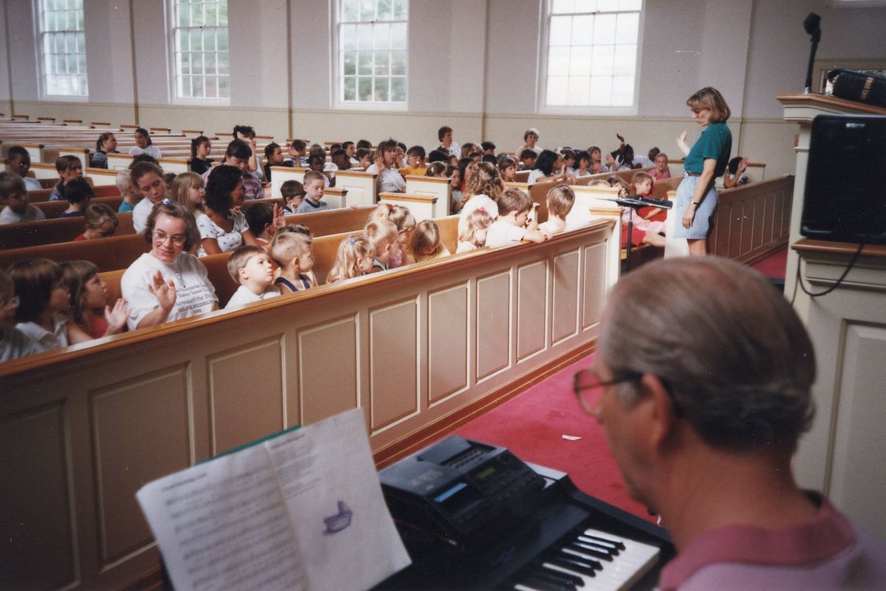 A man plays piano, leading a group of children and a woman in a light-filled room.