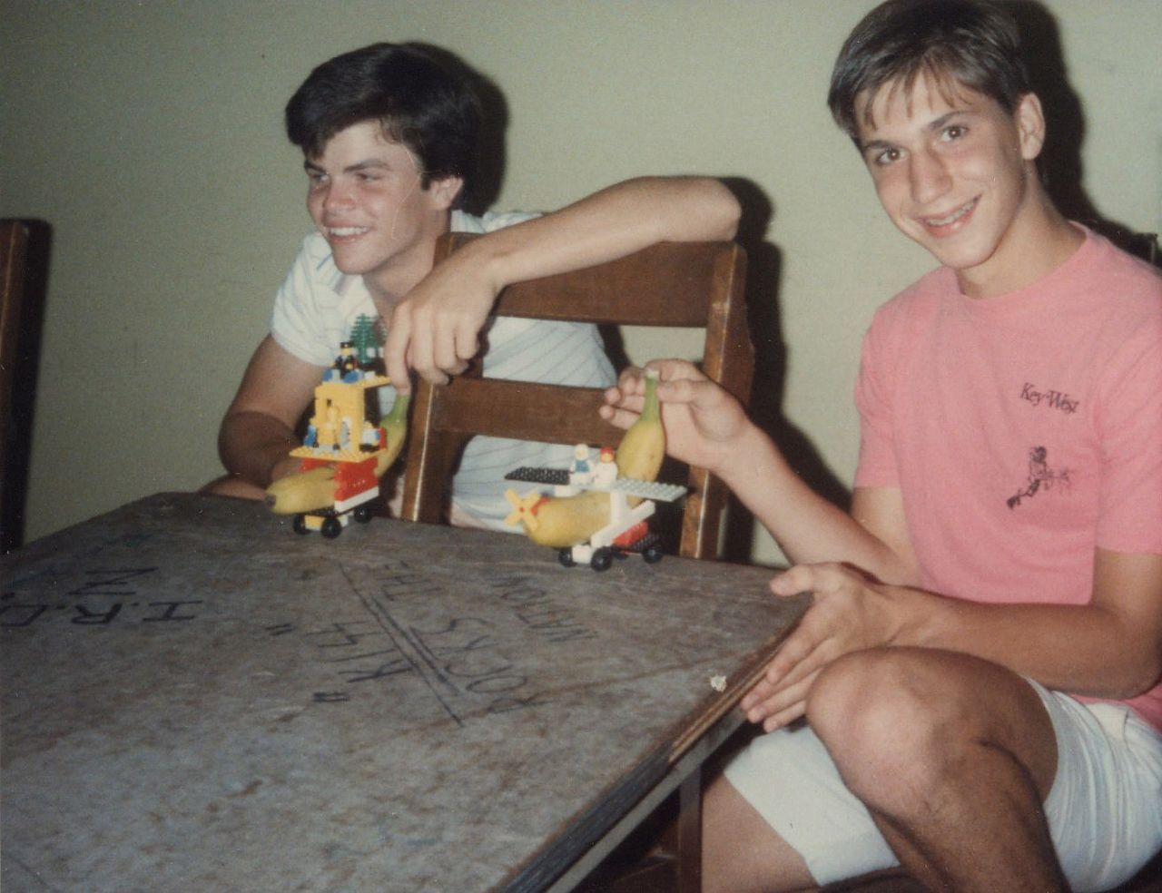 Two boys at a table, smiling and holding toy vehicles. One wears a pink shirt.
