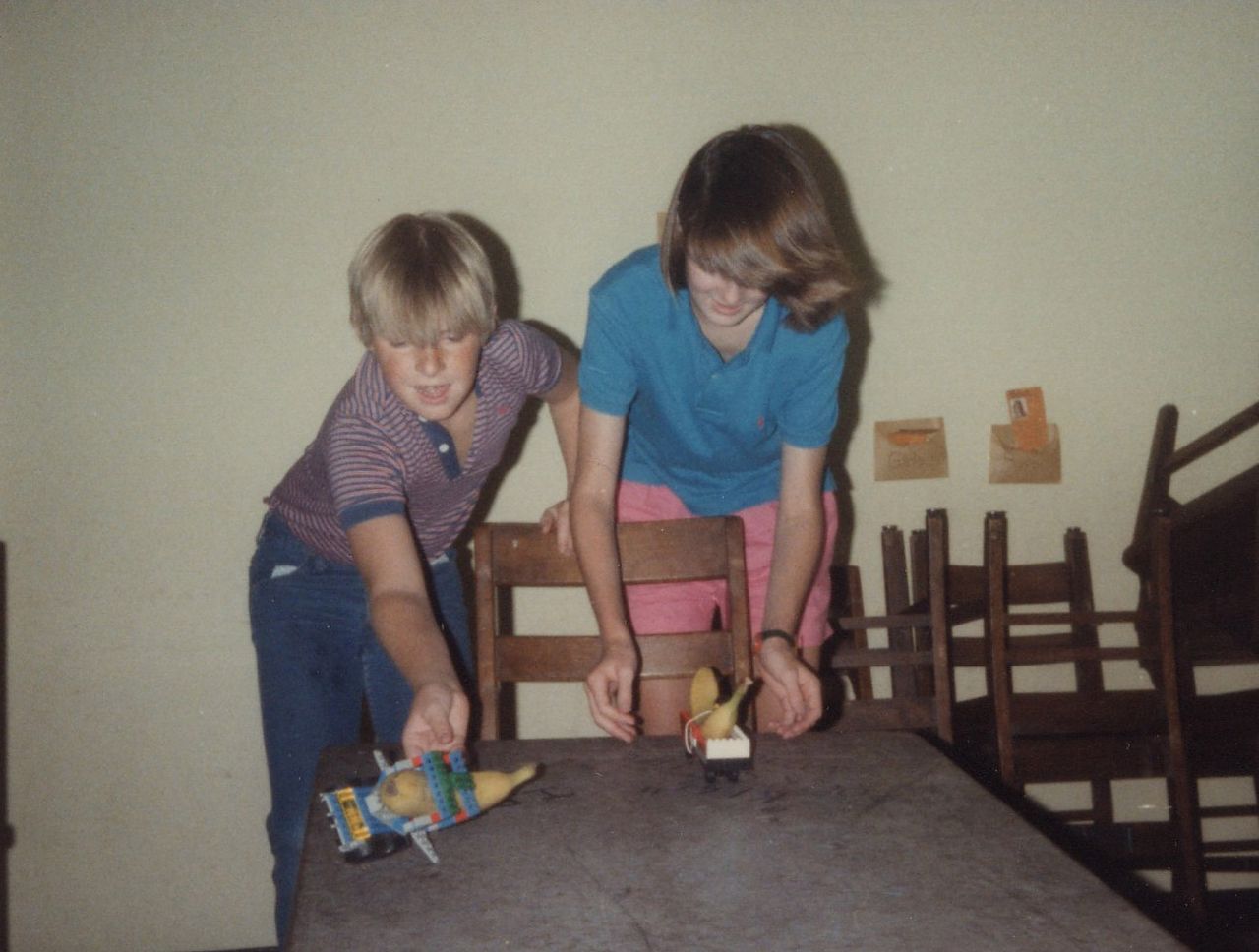 Two people playing with toy cars on a table indoors. One wears a blue shirt, the other a striped shirt.