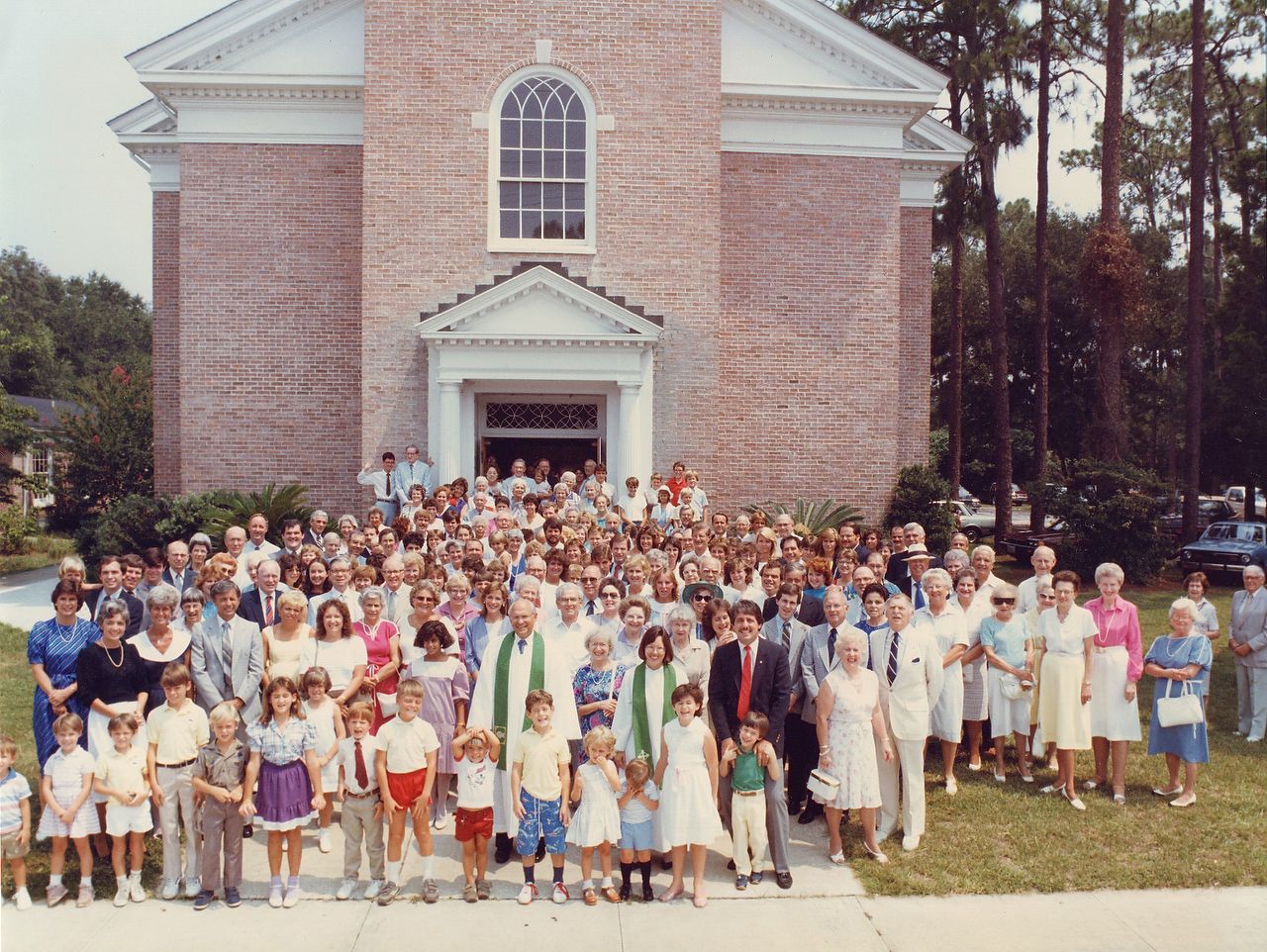 Large group of people pose in front of a brick church with open doors; mostly dressed in light colors.