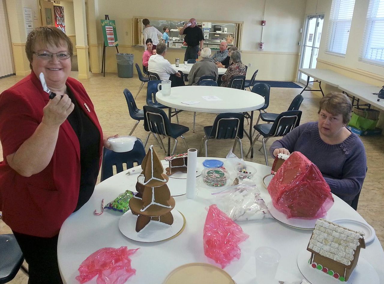 Two women decorating gingerbread houses at a community event. People seated at tables in the background.