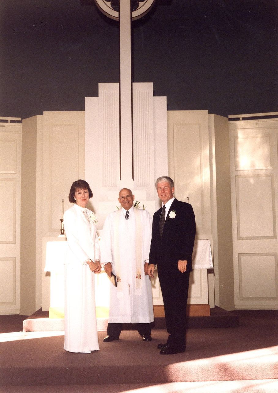 Bride and groom with a priest in a church, standing before a large cross.